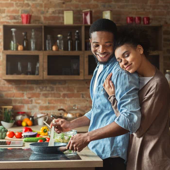 A husband is cooking dinner while his wife hugs him from behind.