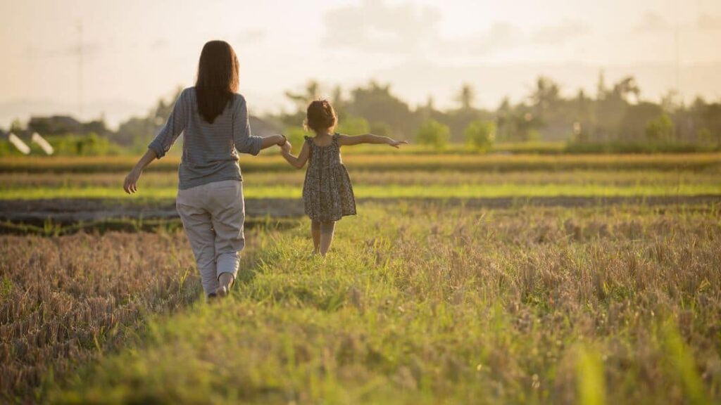 A woman and a young girl walk hand in hand through a grassy field at sunset, enjoying one of the many summer activities for kids. The girl's arms are outstretched as they head toward the horizon, surrounded by a calm and warm atmosphere.