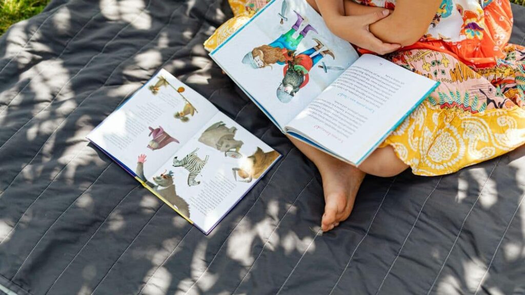 A child in a colorful dress sits on a gray blanket outdoors, enjoying summer activities for kids by reading an illustrated book, with another open picture book nearby. Sunlight and shadows create patterns on the blanket.