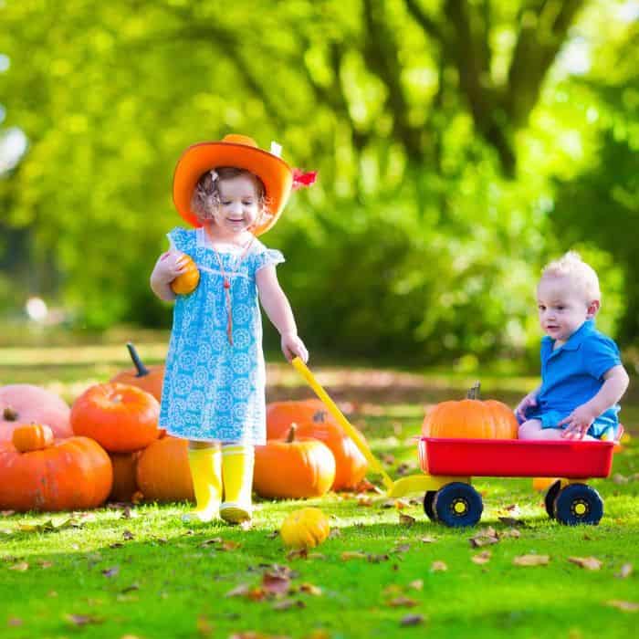 Little girl holding a pumpkin as she pulls her baby brother on a red wagon.