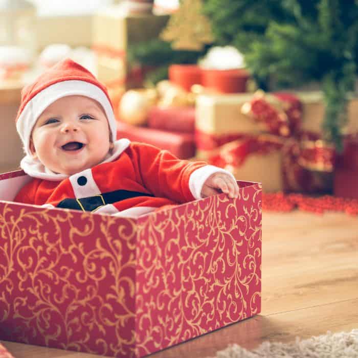 A baby dressed up as Santa Claus sitting in a Christmas present next to a Christmas tree.