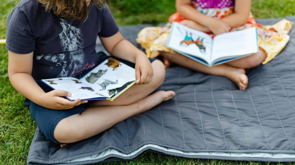 Two children sit cross-legged on a gray blanket outdoors, each reading a book. Enjoying summer activities for kids, one wears a dark shirt and shorts, the other a colorful dress. Both books feature animal pictures.