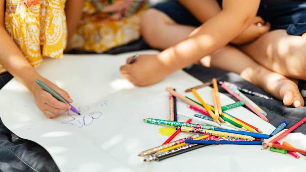 Two children sit on the ground, drawing on a large white paper with colored pencils and markers scattered around them—one of the perfect summer activities for kids. Only their hands and legs are visible as sunlight filters through, casting dappled shadows.