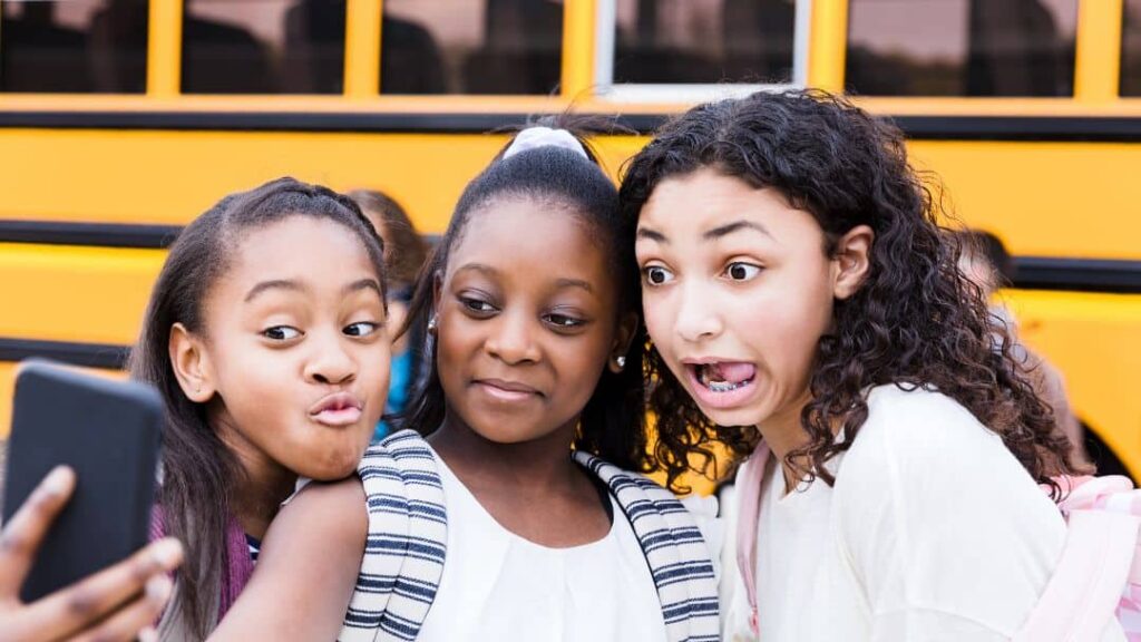 Three school-aged girls with backpacks take a selfie in front of a yellow school bus, each making different playful facial expressions—capturing the fun spirit of great LDS activity day ideas for girls.