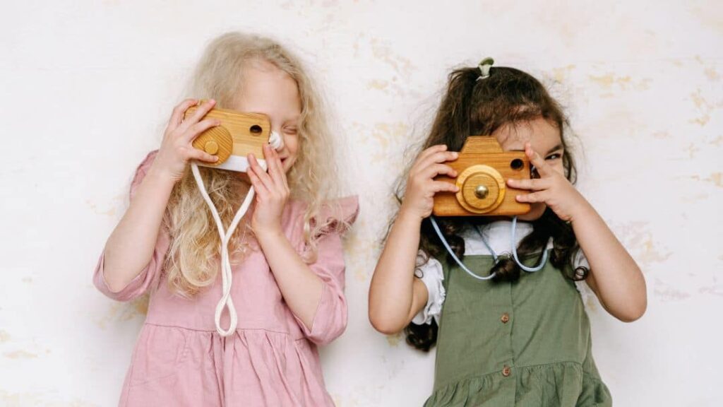 Two young girls stand side by side, smiling and pretending to take photos with wooden toy cameras—an adorable scene and a fun inspiration for LDS activity day ideas for girls. Both wear colorful dresses and pose against a light background.