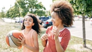 Two young people smile and play basketball outdoors on a sunny day, with trees and parked cars in the background—perfect inspiration for LDS activity day ideas for girls who enjoy fun and active games together.