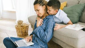 A woman sits on the floor using a laptop while a smiling young boy leans over the couch to hug her. A wicker basket with a teddy bear and an open book are nearby in a bright living room.
