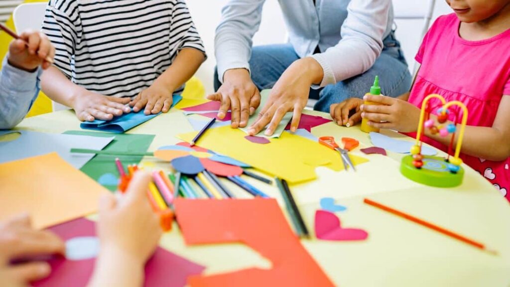 Several children and an adult sit around a table doing arts and crafts with colored paper, pencils, glue, and small toys—hands busy cutting, folding, and gluing. It's a fun example of LDS activity day ideas for girls to encourage creativity.