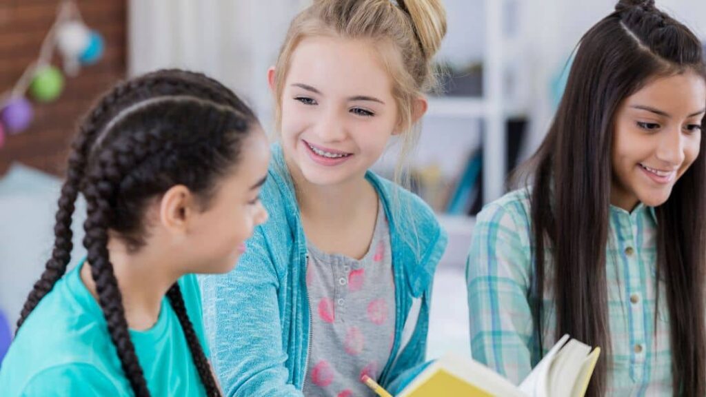 Three smiling girls sit close together, reading a book and chatting. They appear to be friends enjoying each other’s company, sharing fun LDS activity day ideas for girls in a bright, indoor setting.