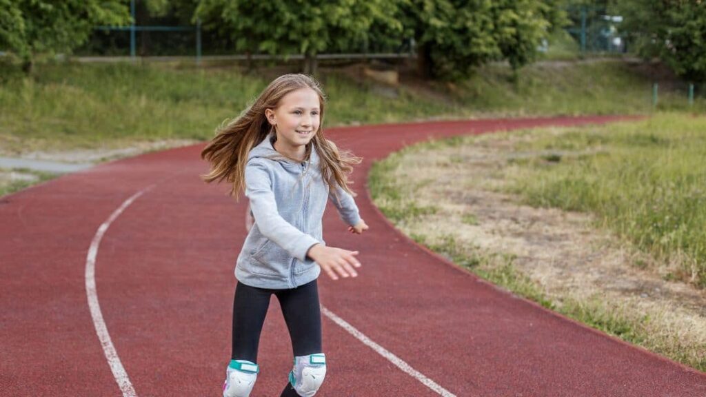 A young girl with long hair rollerblades on a red outdoor track, wearing a gray hoodie, black leggings, and knee pads. She is smiling, surrounded by greenery—perfect inspiration for LDS activity day ideas for girls.