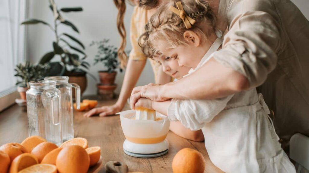 A woman helps a young child squeeze fresh orange juice into a juicer on a wooden kitchen counter, surrounded by whole and halved oranges, glass pitchers, and potted plants—one of many fun summer activities for kids.