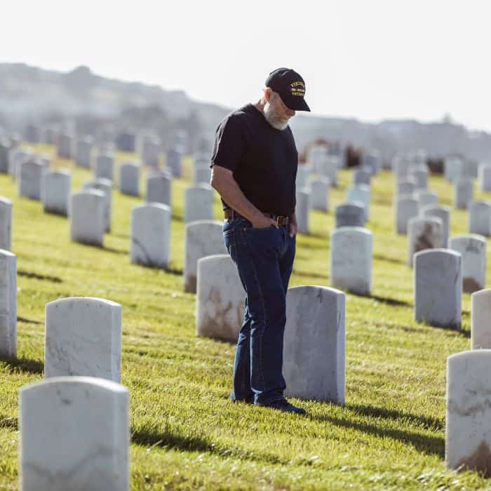 A man in a black shirt and cap standing solemnly at a grave in a cemetery with numerous white headstones in the background, reflecting a moment of remembrance and respect.
