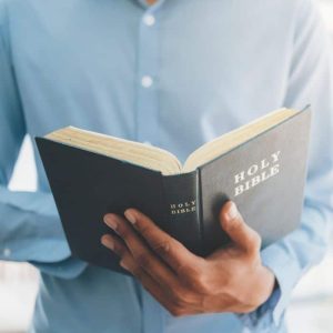 A man in a light blue shirt holds an open holy bible, reading it intently. the focus is on the book and the person's hands.