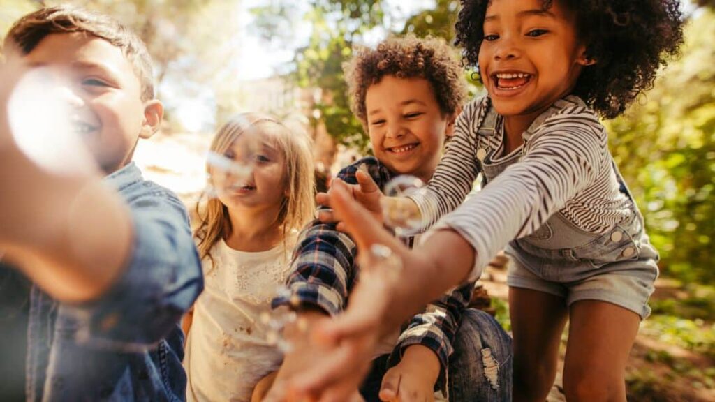Four smiling children reach out excitedly towards floating bubbles outdoors, with sunlight filtering through trees in the background—a perfect example of summer activities for kids as they play joyfully together.