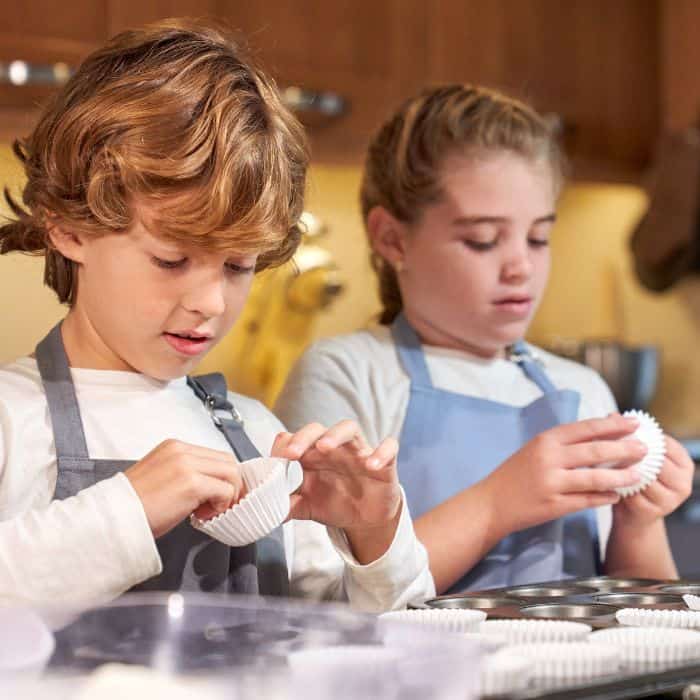 Two children, a boy and a girl, are intently preparing cupcake liners in a kitchen, both wearing aprons. The setting suggests an activity day baking session.