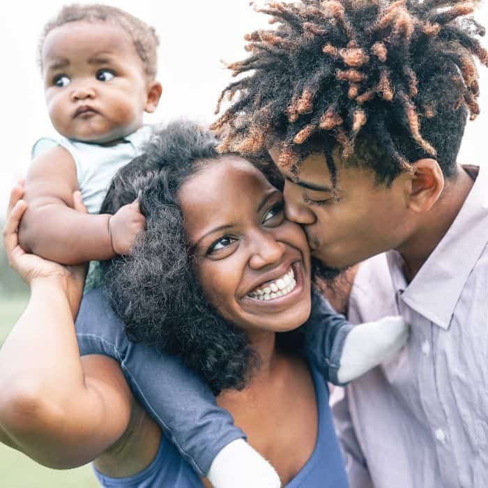 An older son kissing his mom's cheek, while the mother balances her new baby boy on her back.