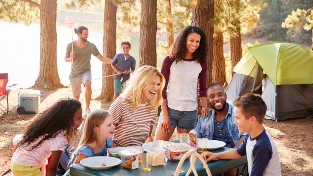 A group of people, including adults and children, gather around a picnic table with food, smiling and laughing at a campsite among trees—a perfect scene for fun summer ideas with friends, with tents and camping gear in the background.