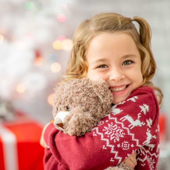 A little girl holding a teddy bear in front of the christmas tree.