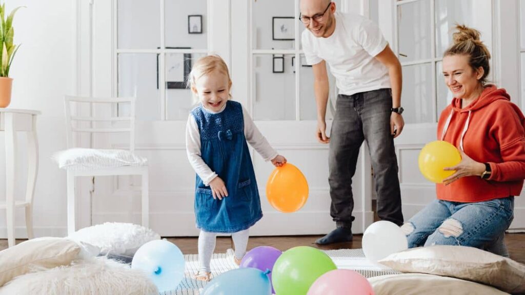 A young child plays with colorful balloons on the floor while two adults smile and watch in a bright, cozy living room with large windows and cushions.