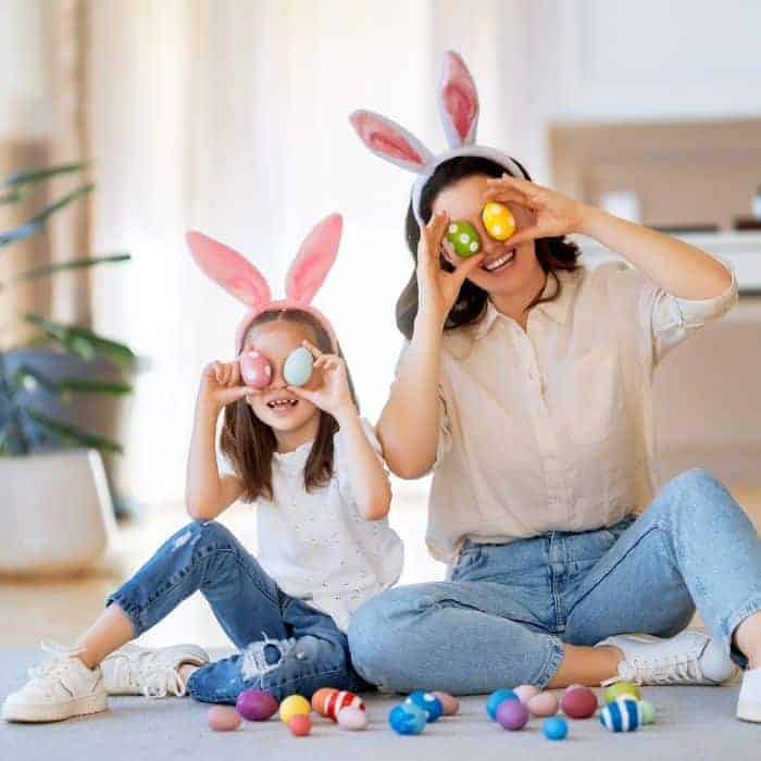 A mother and daughter are wearing Easter bunny headbands and are surrounded by Easter eggs. They are holding a handful of Easter eggs up to their faces.