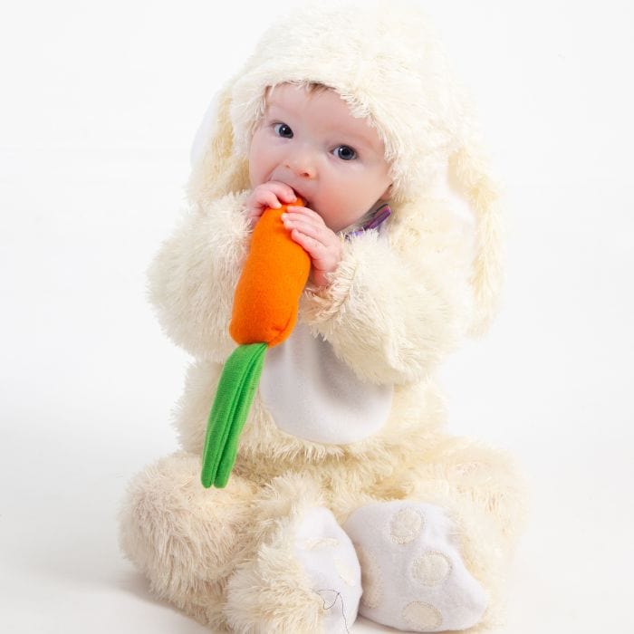 A toddler eating a chew toy in the shape of a carrot while dressed as the Easter bunny.