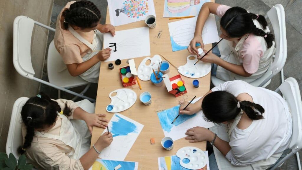 Four children sit around a table painting and drawing on paper, using paint palettes, brushes, and colored markers—perfect LDS activity day ideas for girls. Art supplies and cups are scattered as they work creatively together.