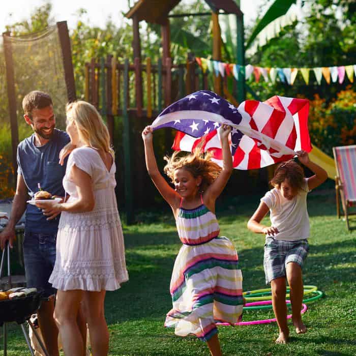 A little girl and boy running around with an American flag at a family bbq.