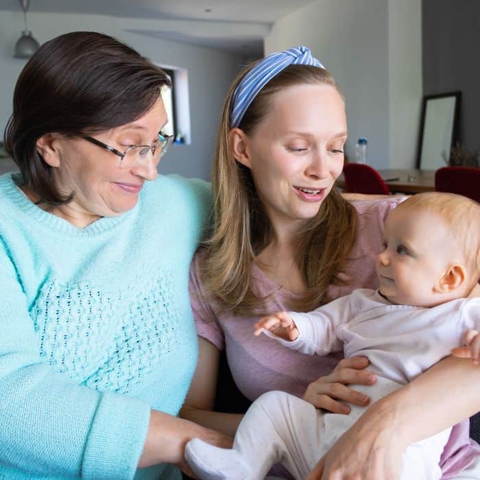 Three generations together: a smiling grandmother, her daughter celebrating her first Mother's Day, and a curious baby share a warm, family moment at home.