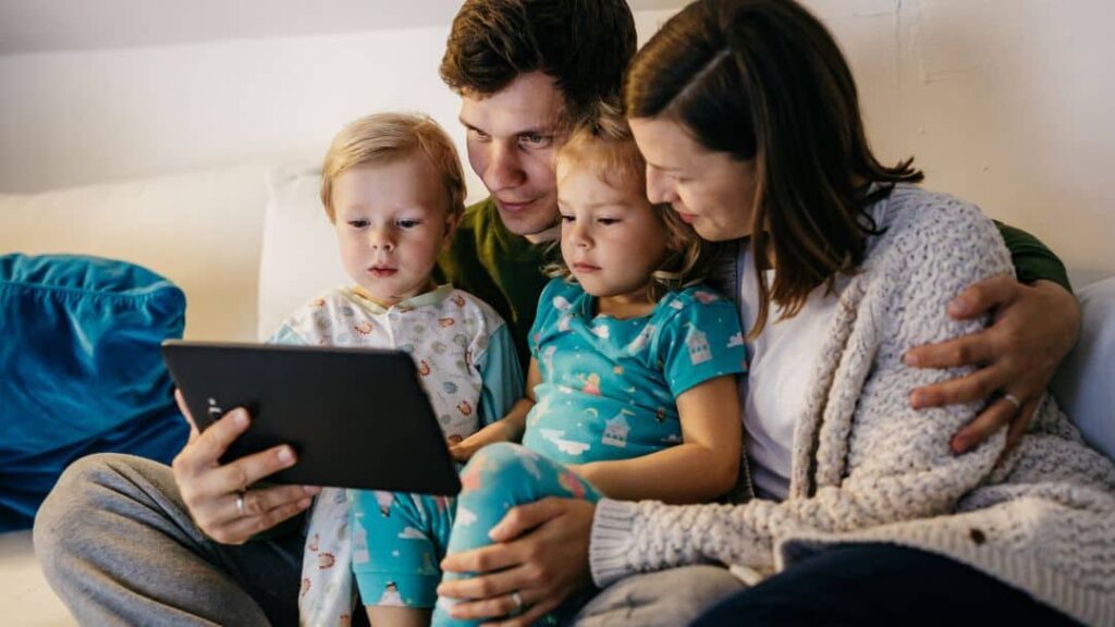 A family of four, two adults and two young children in pajamas, sit closely together on a couch looking at a tablet, appearing engaged and relaxed.