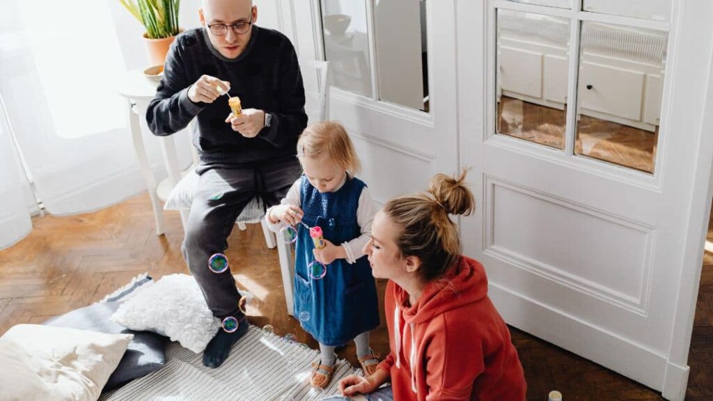 A man, a woman, and a young child blow soap bubbles together while sitting on the floor in a bright, cozy room with wooden floors and white walls.