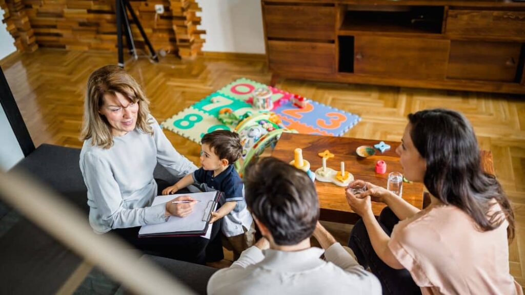 A woman with a notepad talks to a couple and their young child in a living room. Colorful foam mats and toys are on the floor, suggesting a child-friendly environment. The adults appear engaged in discussion.