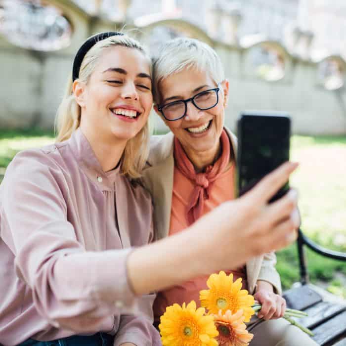 An older and younger woman sitting next to each other, smiling. One is holding her phone as they take a picture.