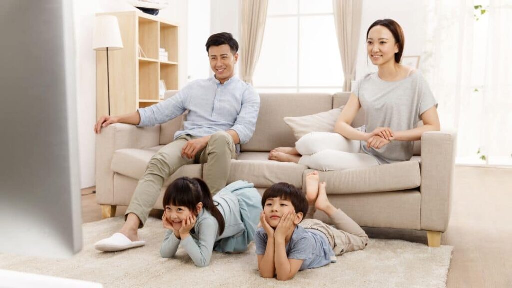 A family of four relaxes together in a living room, with two children lying on the carpet in front of the TV and two adults sitting on the couch behind them, all smiling and watching something.