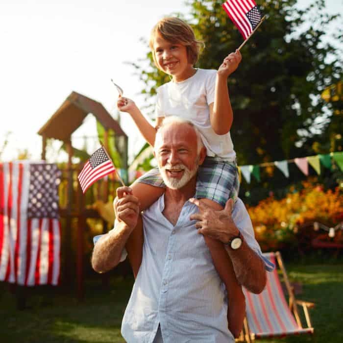 A little boy sitting on his grandpa's shoulders waving an American flag.