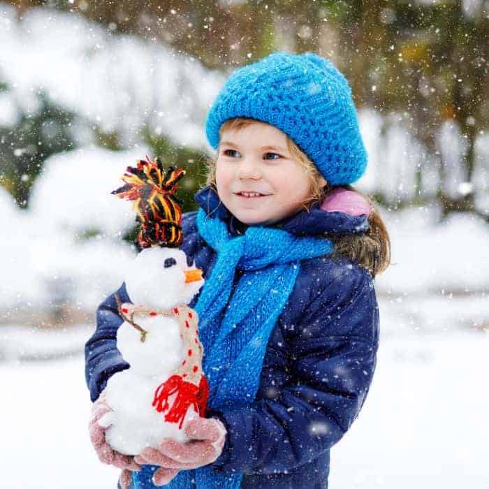 A toddler carrying a small snowman in her hands while playing in the snow outside in her snowsuit.