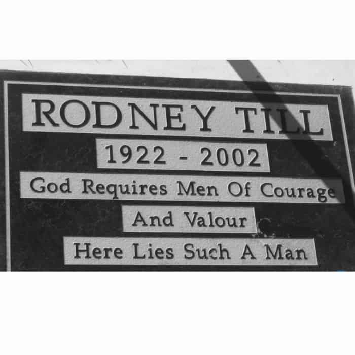 A solemn tribute: a black and white photo of a memorial plaque reading "rodney till 1922 - 2002, god requires men of courage and valour here lies such a man", casting a sharp shadow across its surface.
