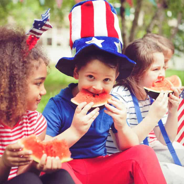 A little boy in an American flag hat eating a slice of watermelon with his friends.