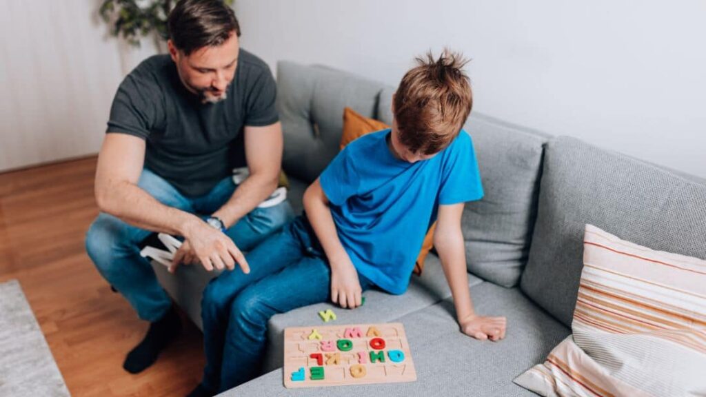 A man and a boy sit on a gray couch. The boy, wearing a blue shirt, plays with a wooden alphabet puzzle while the man, in a dark shirt, points and interacts with him. Pillows are in the background.