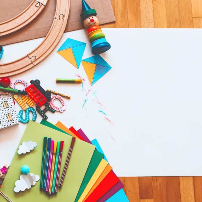 A variety of colorful children's art supplies including markers, colored paper, and toys on a wooden floor with a blank white sheet ready for crafting.