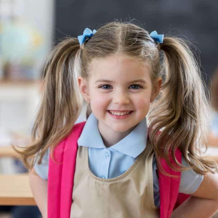 A young girl with pigtails and blue bows smiling at the camera, wearing a pink backpack and a school uniform with a blue shirt and beige jumper in a classroom.