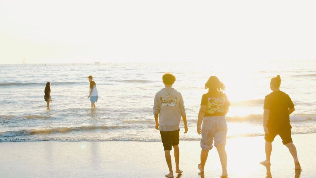 Four people walk toward the ocean on a sunny beach, with two others standing in the shallow water. The bright sunlight and relaxed atmosphere show one of many fun summer ideas with friends.