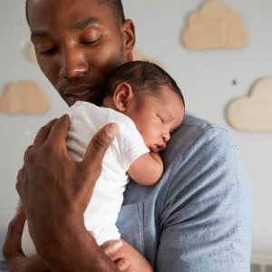A man gently holds a sleeping baby close to his chest. The man wears a light blue shirt, and the baby is dressed in a white onesie. The background features wall decorations shaped like clouds, exuding warmth and tenderness akin to LDS Father's Day quotes celebrating love and family.