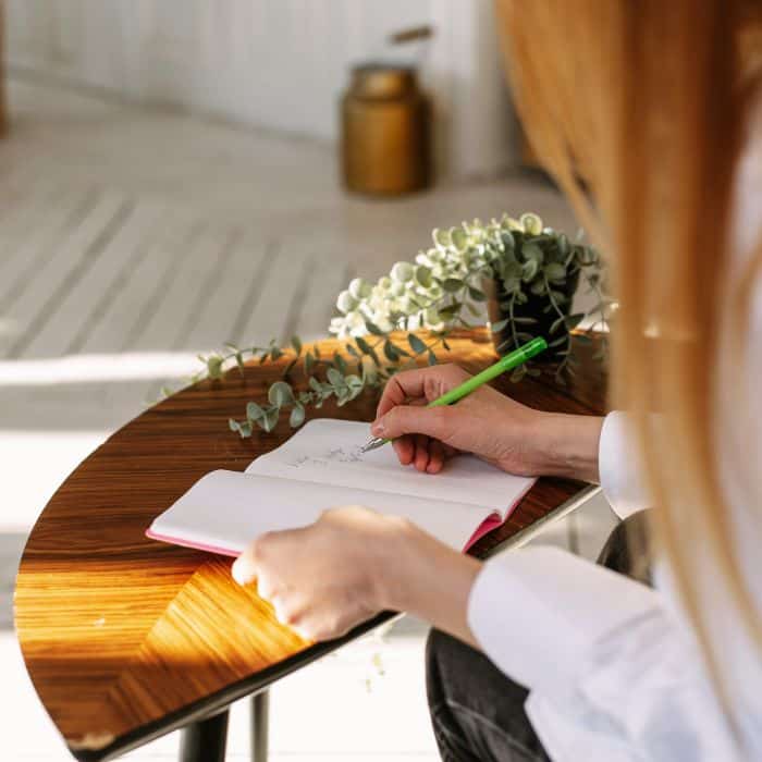 A person is sitting at a wooden table, writing or sketching in a mental health printable journal with a pencil, alongside a decorative plant. Sunlight streams in, creating a warm, bright workspace.