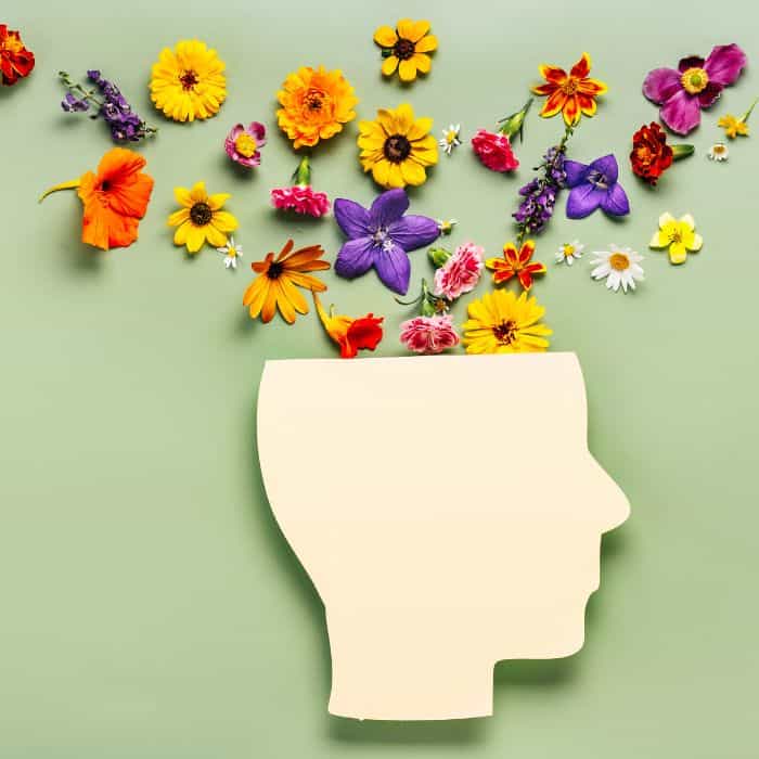 A creative display featuring a silhouette of a human head outlined with yellow paper on a light green background, surrounded by a burst of colorful, assorted flowers, representing mental health themes in printable journal prompts.