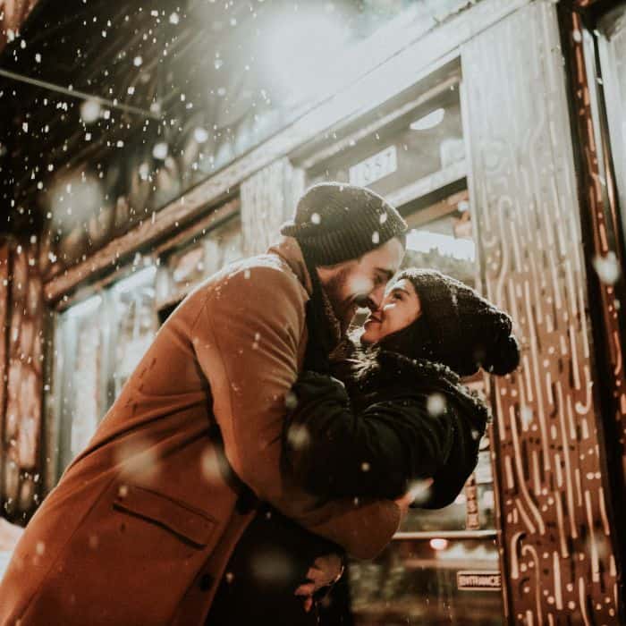 A happy couple caught in the snow at night. Both are wrapped up together about to kiss.