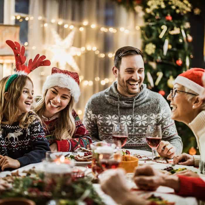 A family sitting around the dinner table with Christmas hats and lights. There's food on the table and everyone is smiling and laughing.