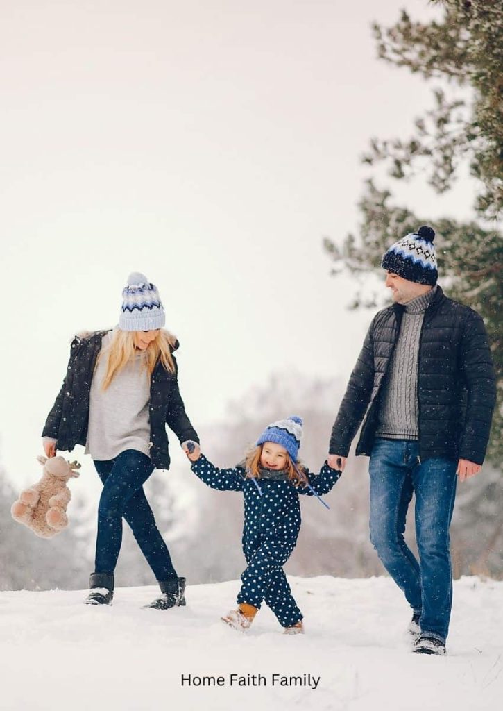 A young family holding hands and walking in the snow. The mom is holding a teddy bear.