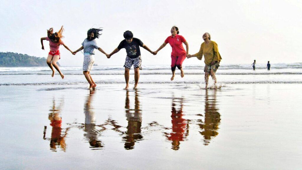 Five people holding hands and jumping on a beach, their reflections visible on the wet sand—perfect for fun summer ideas with friends. The ocean and a few distant people are in the background under a clear sky.