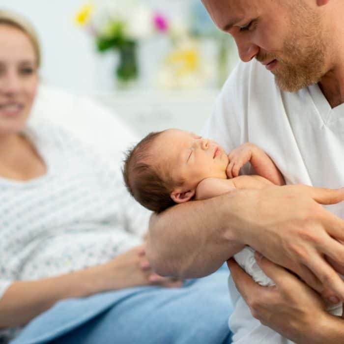 Dad holding newborn with mom laying in hospital bed.