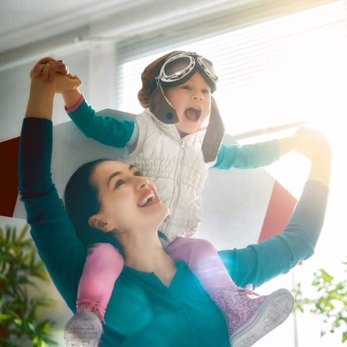 A joyful moment as a mother lifts up her child, who is wearing pilot goggles and a scarf, pretending to fly with excitement and imagination.